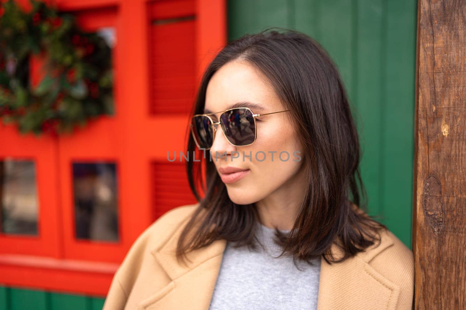 People, season and Christmas concept portrait of happy smiling middle age woman outdoors dressed stylish cream trench coat with Christmas festive decoration on background. Looking away, copy space