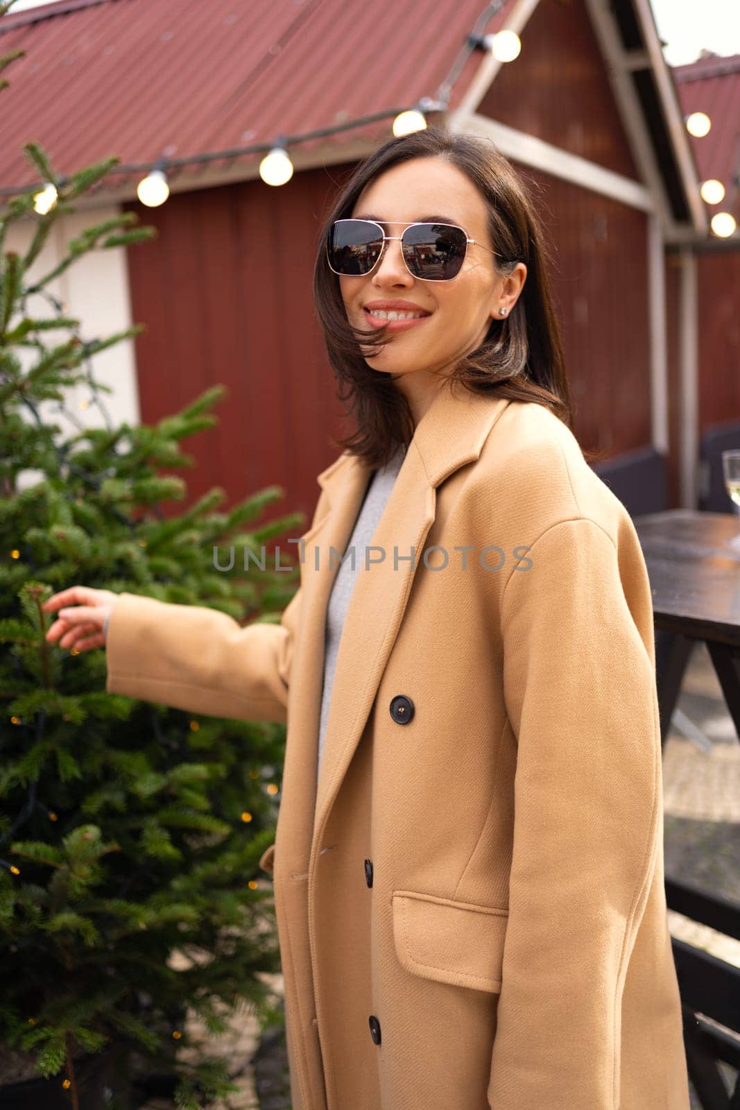 Woman looking for perfect Christmas tree to buy on the tree shop, outdoor in European city. Girl using hand for check the quality. Dressed stylish trench coat and sunglasses
