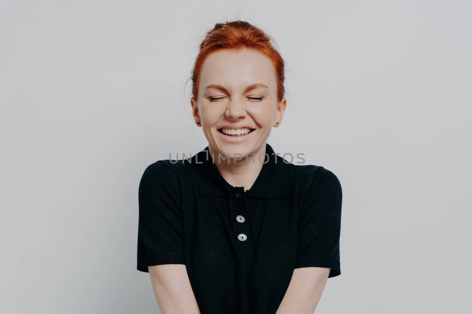 Happy pleased red haired woman with closed eyes and toothy smile posing against grey wall by vkstock