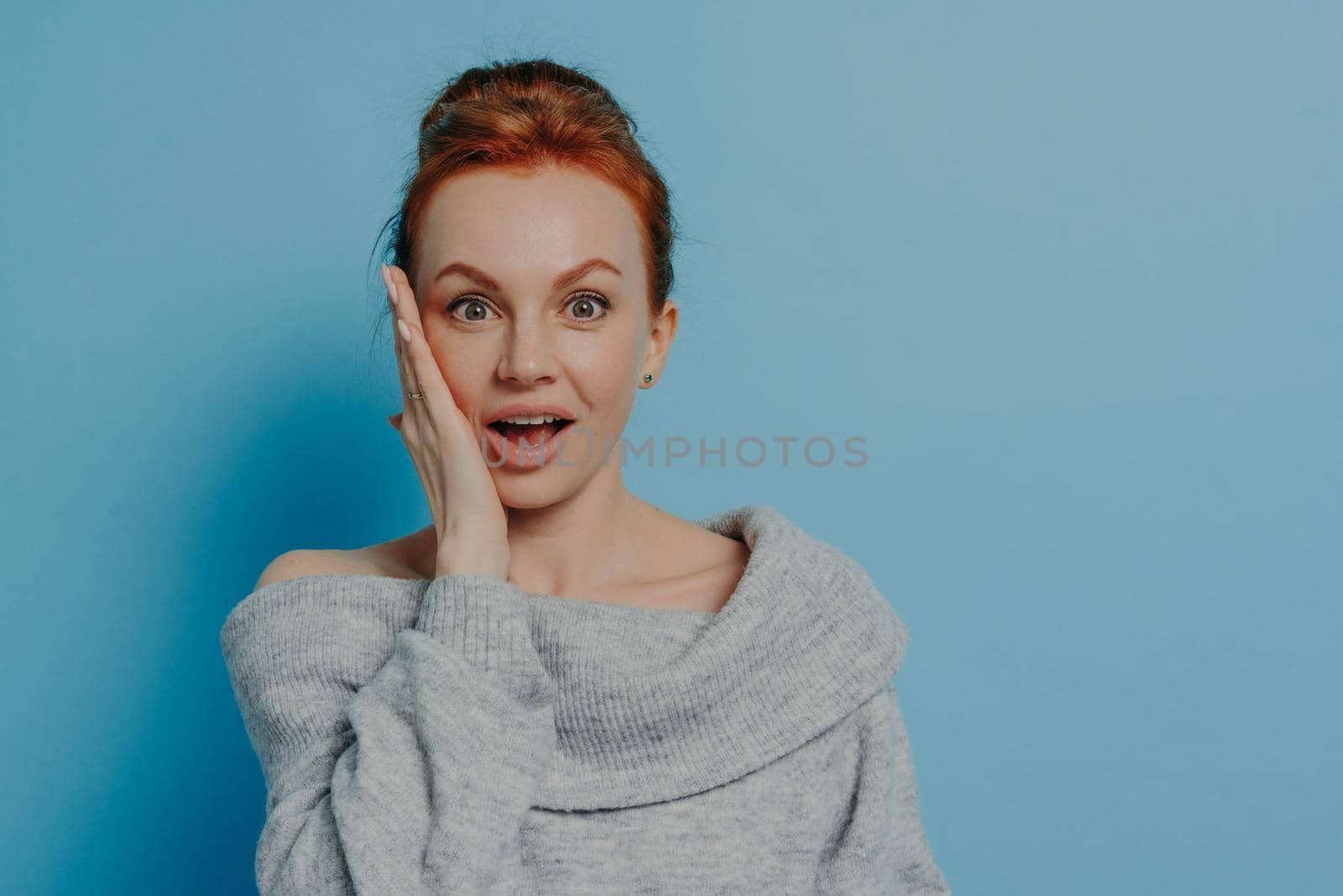 Portrait of surprised amazed red haired woman looking at camera with impressed face expression, feeling happy after hearing unexpected news, isolated on blue studio background, female being shocked