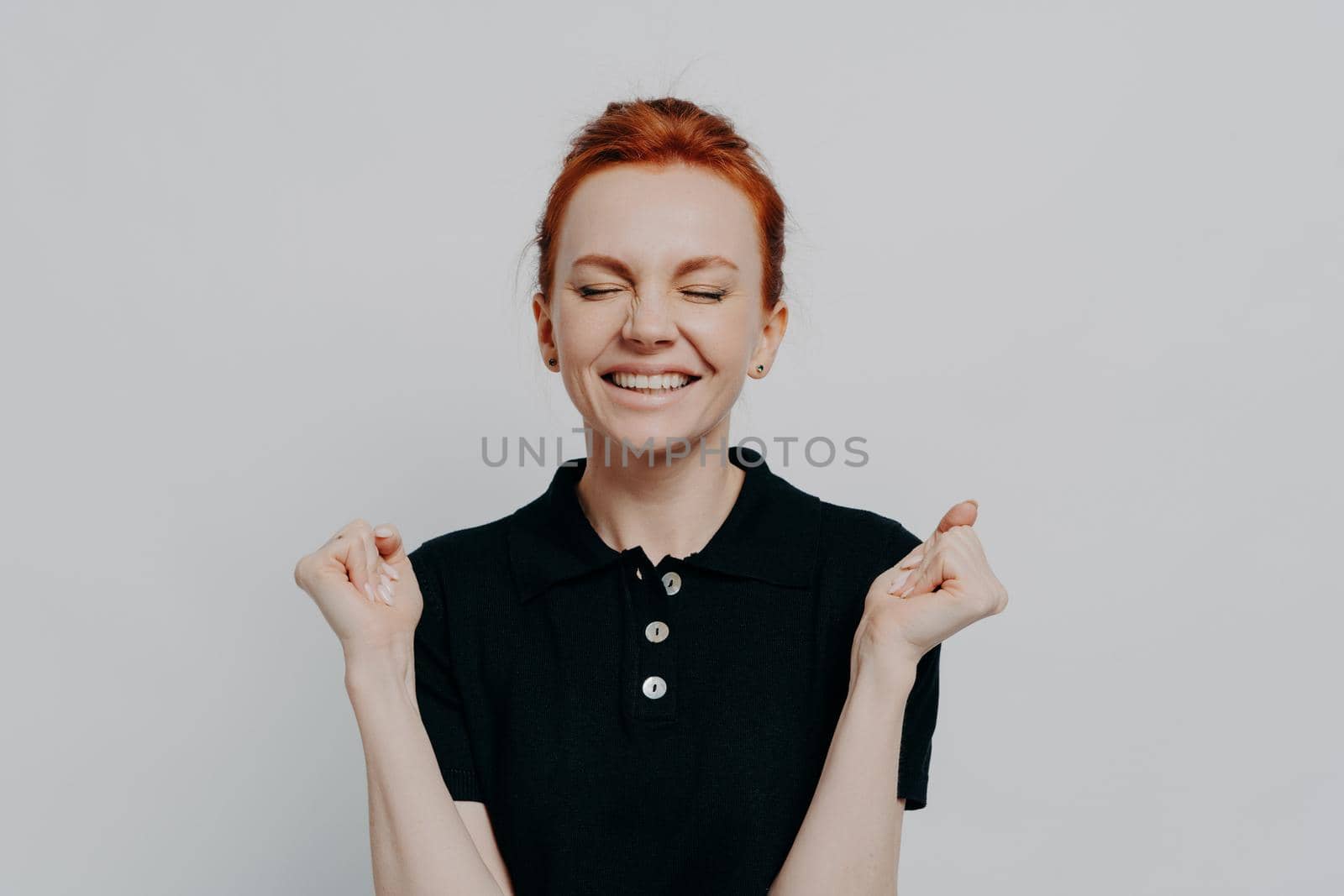 Yes, i did it. Portrait of happy euphoric red haired female with closed eyes holding clenched fists up, excited woman celebrating win, success while posing isolated over grey studio wall
