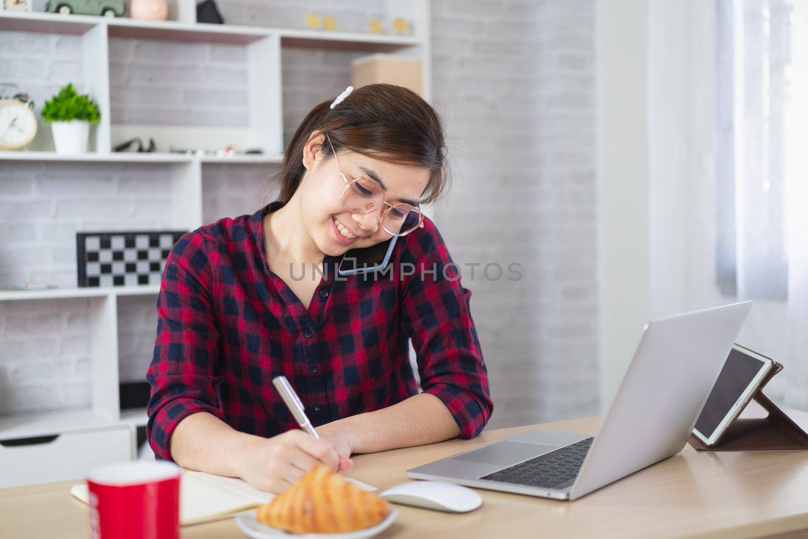 Beautiful asian woman talking on cellphone consulting client or customer online and using laptop computer while sitting on table. by Wmpix