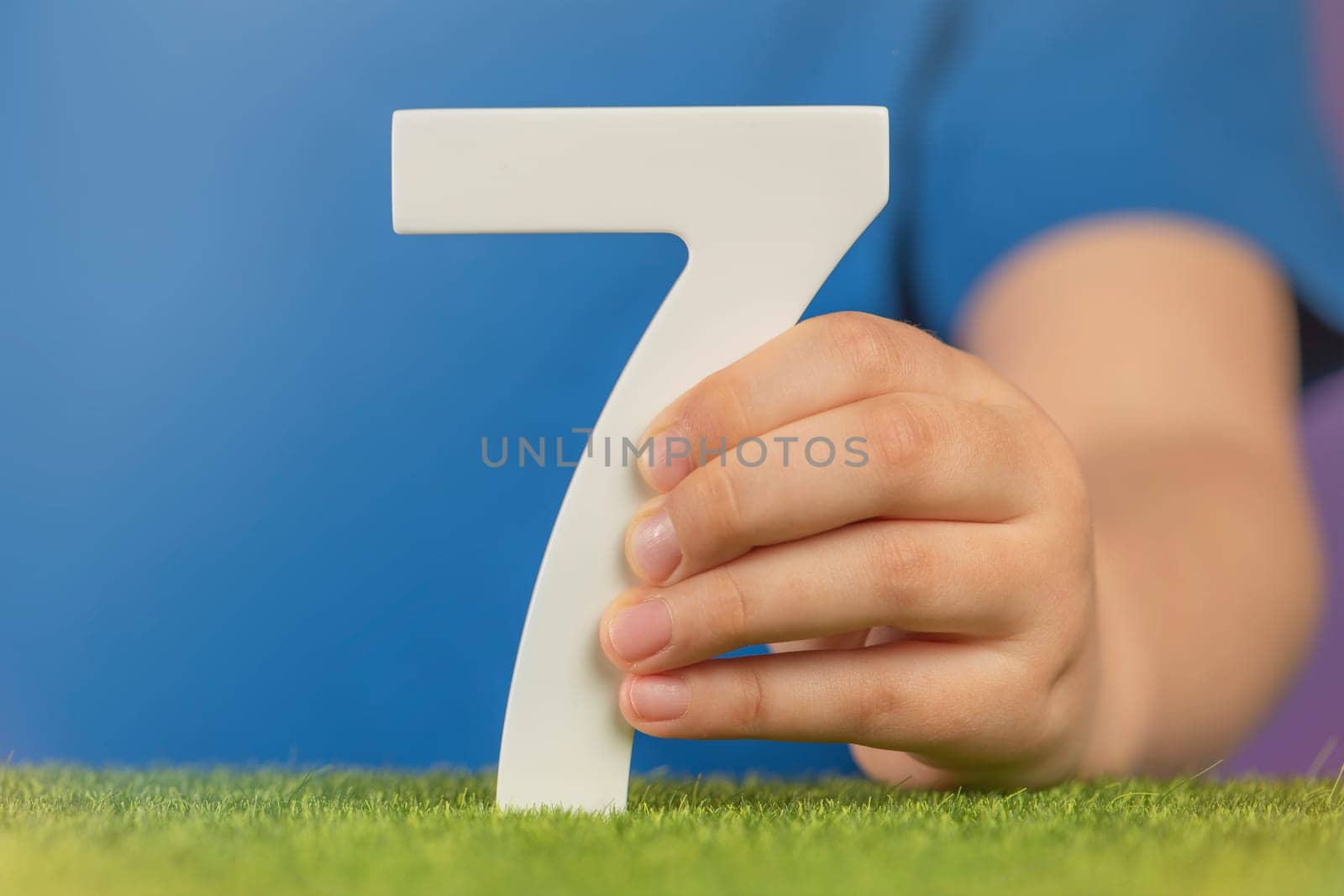 Number 7 in hand. A hand holds a white number seven on green grass and a purple background, seven percent per annum. World Health Day. April 7th. Holiday concept. by SERSOL