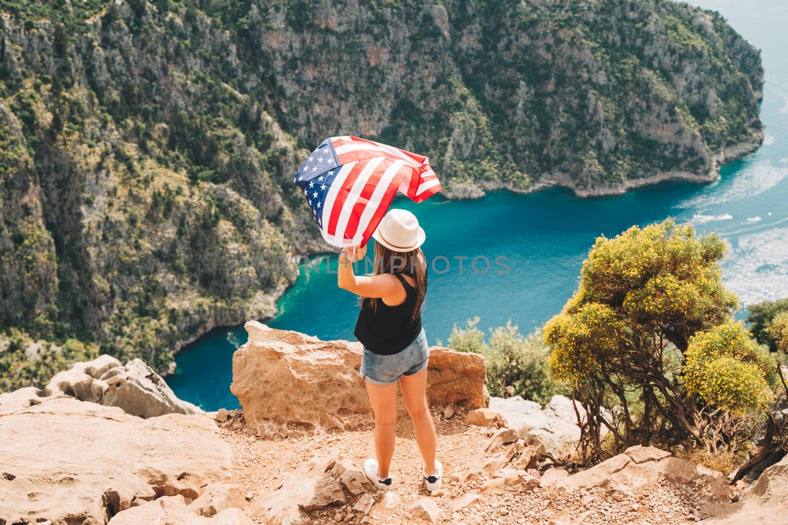 Young woman standing on a rock cliff and waving the US flag while looking at sea beneath. Girl traveller waving the American flag while standing on a mountain top. 4 fourth July Independence Day