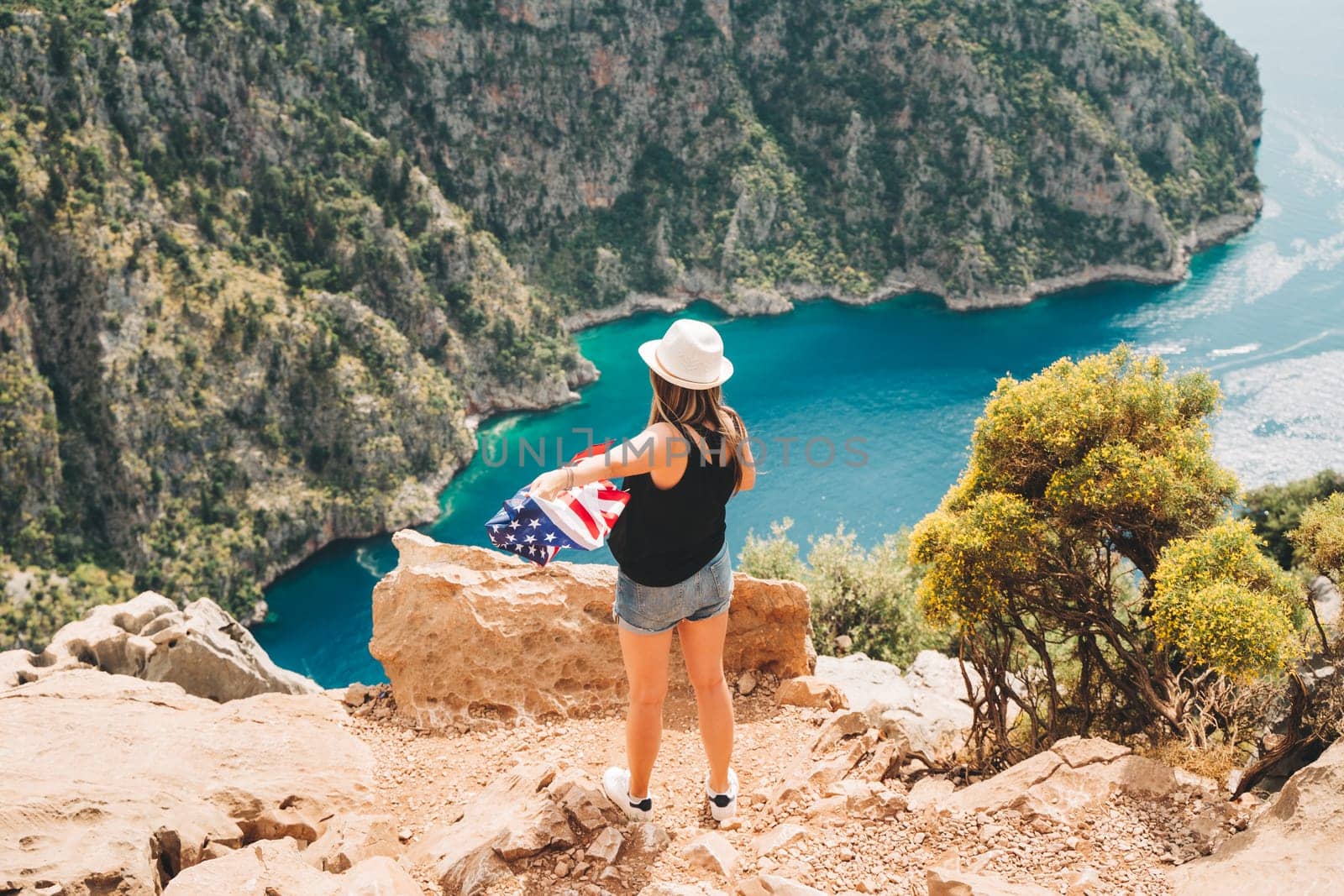Young woman standing on a rock cliff and waving the US flag while looking at sea beneath. Girl traveller waving the American flag while standing on a mountain top. 4 fourth July Independence Day