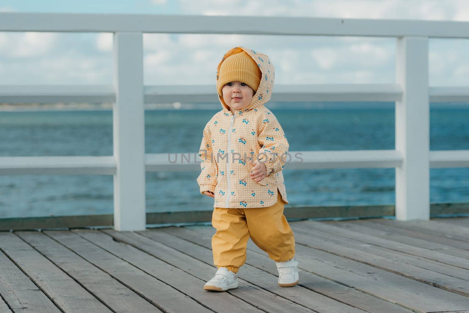 A beautiful toddler in a yellow jacket and pants strolls on the pier. A baby girl in the hood is having fun near the Baltic Sea.