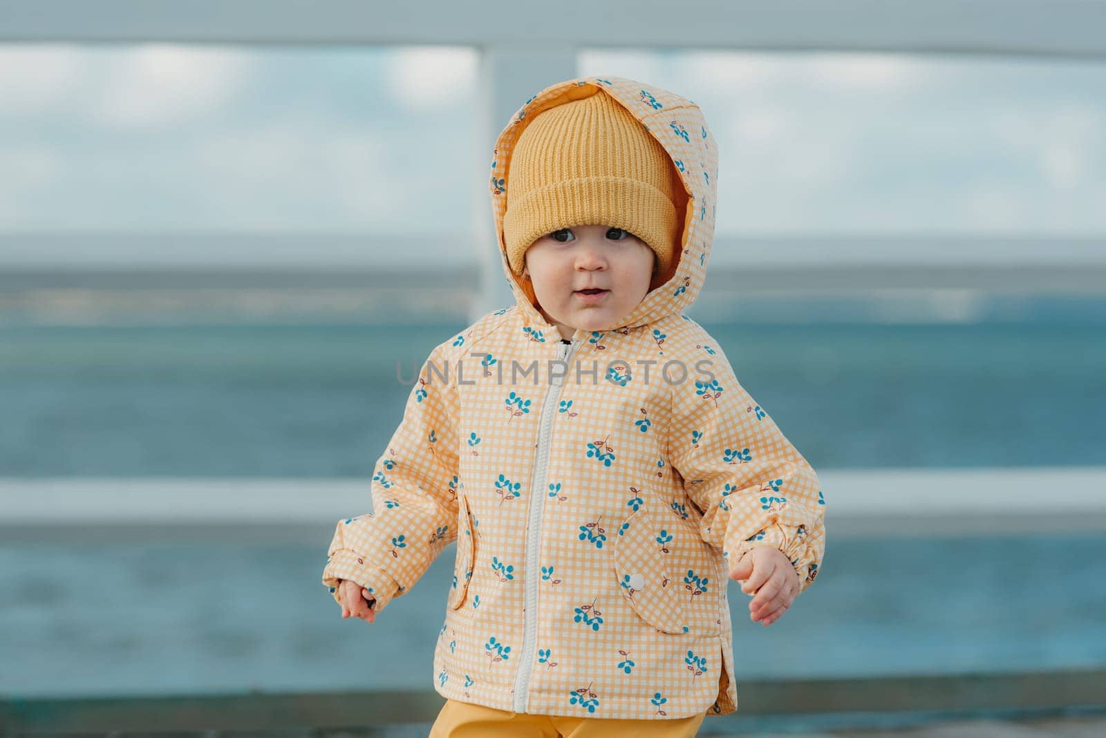 A curious toddler in a yellow jacket and pants strolls on the pier. A baby girl in the hood is having fun near the Baltic Sea.