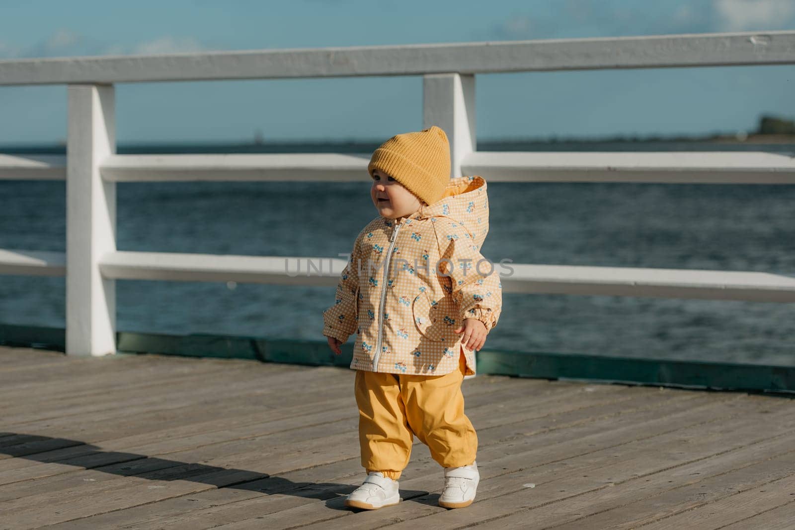 A toddler in a yellow jacket and pants walks on the pier by RomanJRoyce