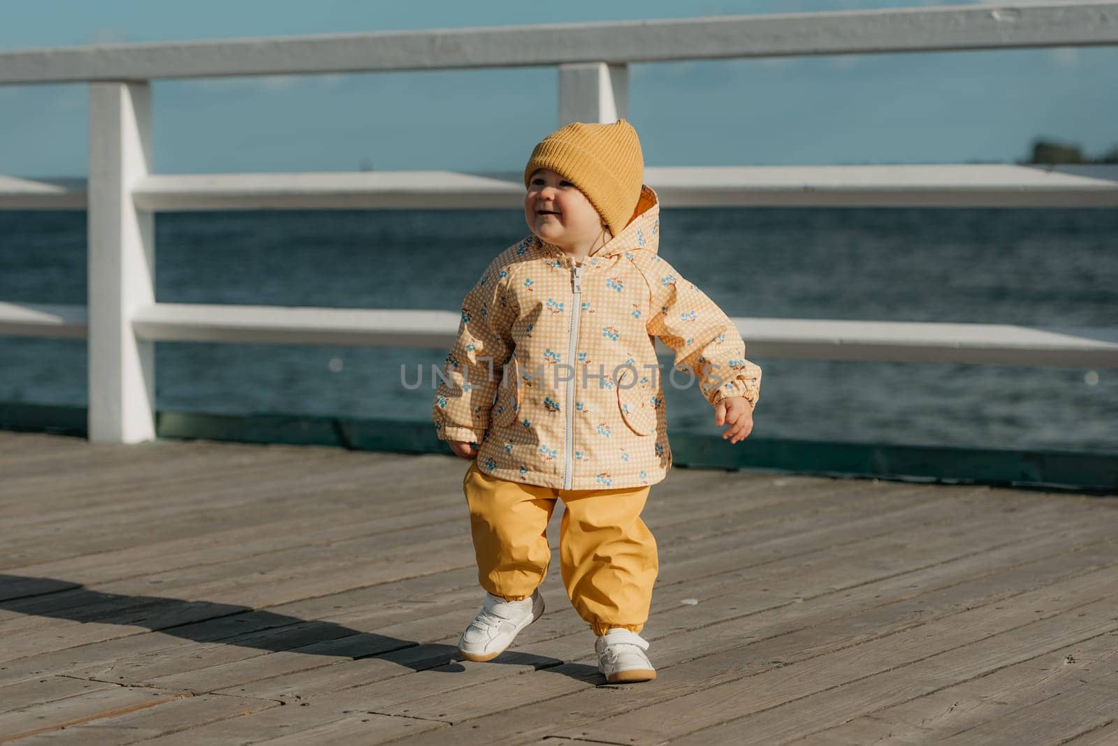 A happy toddler in a yellow jacket and pants strolls on the pier. A baby girl is having fun near the Baltic Sea.