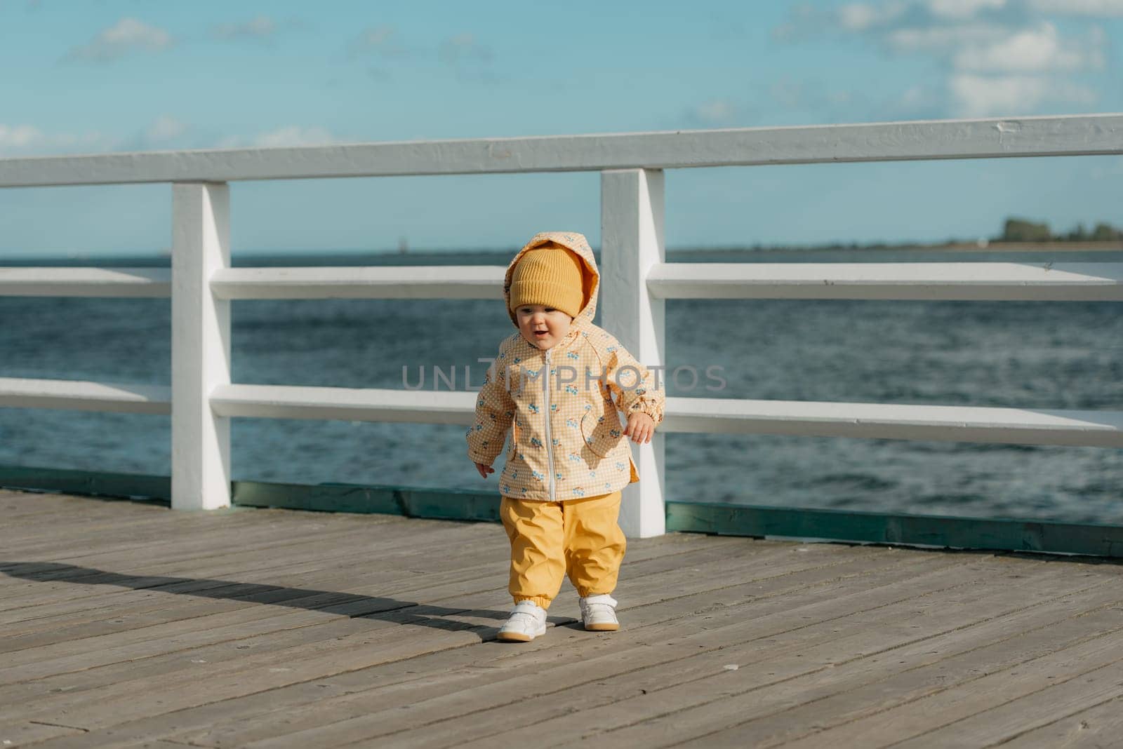 A smiling toddler in a yellow jacket and pants walks on the pier. A baby girl is having fun and screaming near the Baltic Sea.