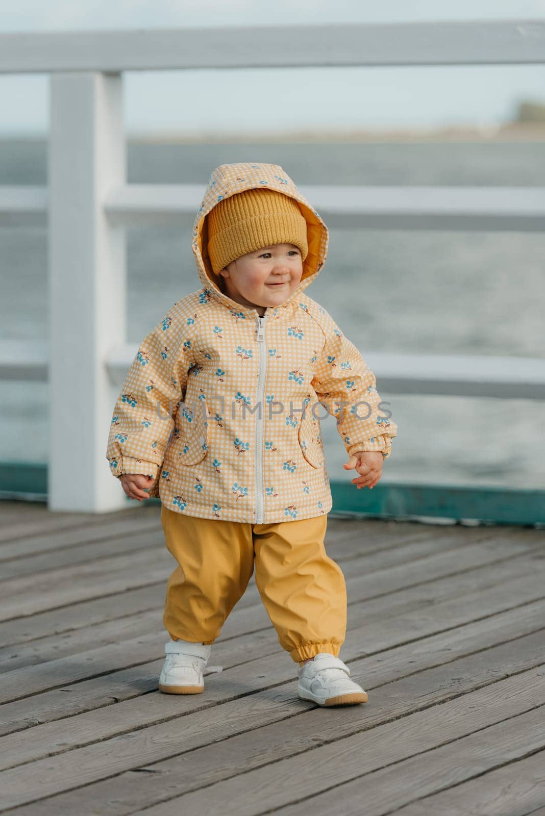 A smiling toddler in a yellow jacket and pants walks on the pier. A baby girl is having fun near the Baltic Sea.