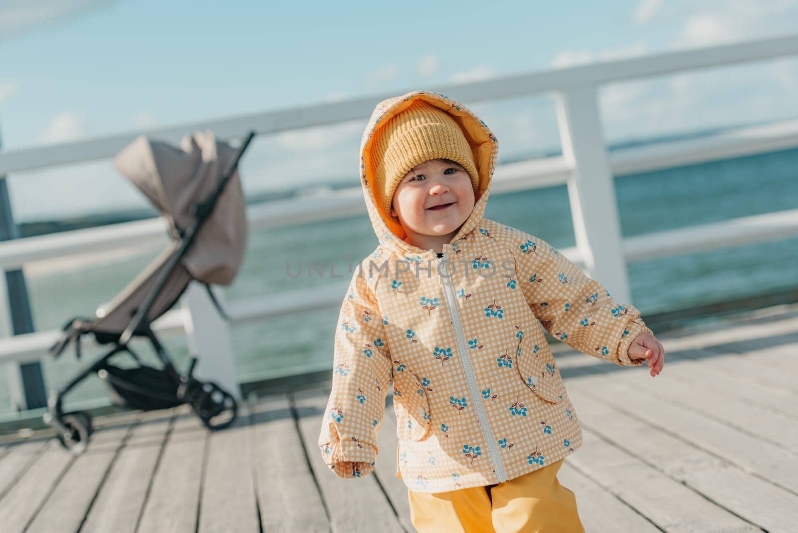 A toddler in a yellow jacket and pants walks on the pier by RomanJRoyce