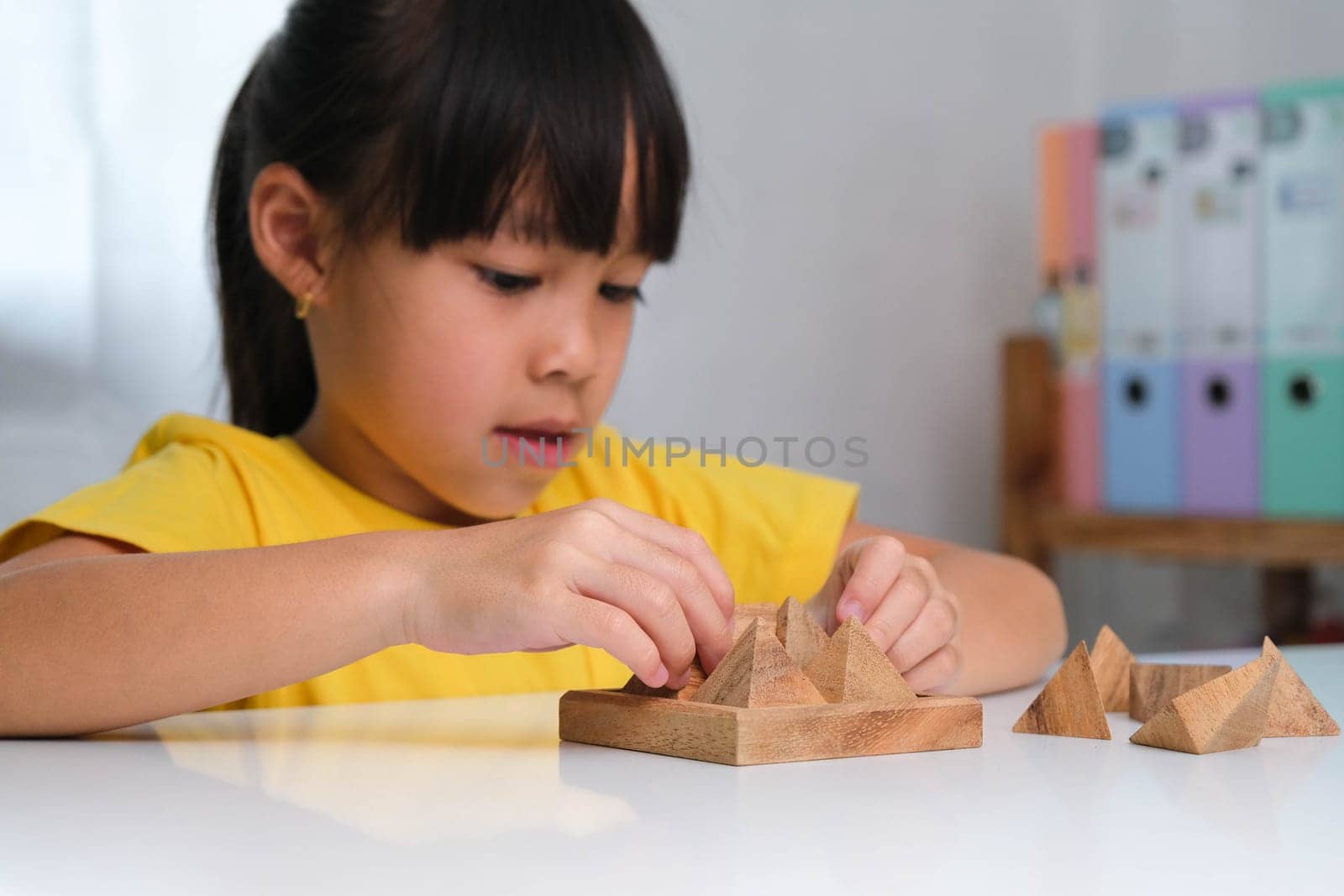 Asian cute little girl playing with wooden toy jigsaw puzzle pyramid on table. Healthy children training memory and thinking. Wooden puzzles are games that increase intelligence for children. Educational toys for children. by TEERASAK