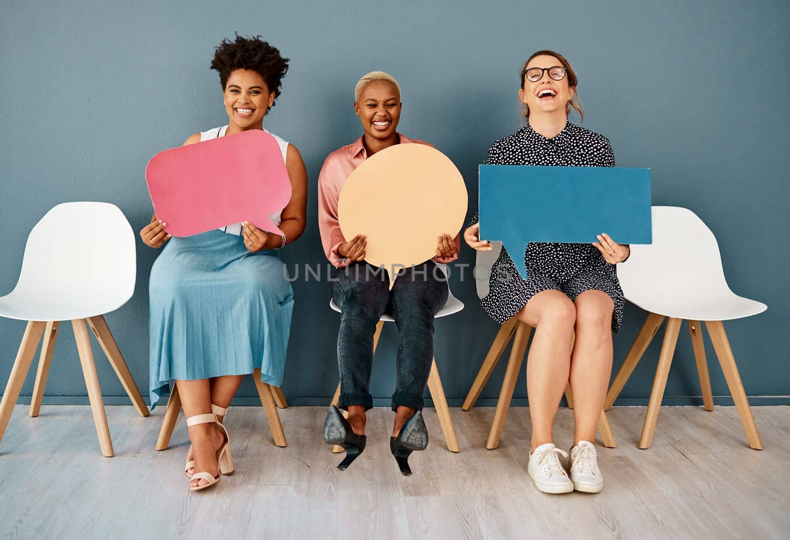 Isnt that just funny. Studio portrait of a group of attractive young businesswomen holding speech bubbles while sitting in a row against a grey background. by YuriArcurs