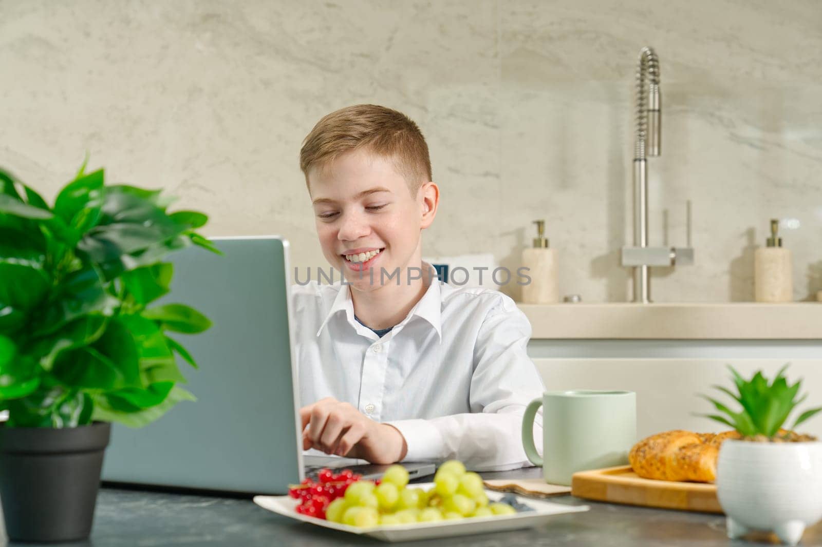 young boy, teenager plays on the notebook during breakfast time. Addiction to gadgets. Breakfast before school in the kitchen.