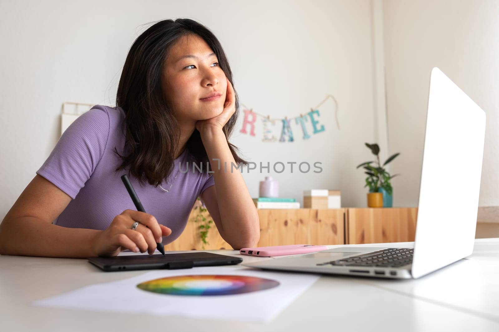 Asian young female graphic design student working on a project at home using tablet and laptop. Color wheel. by Hoverstock