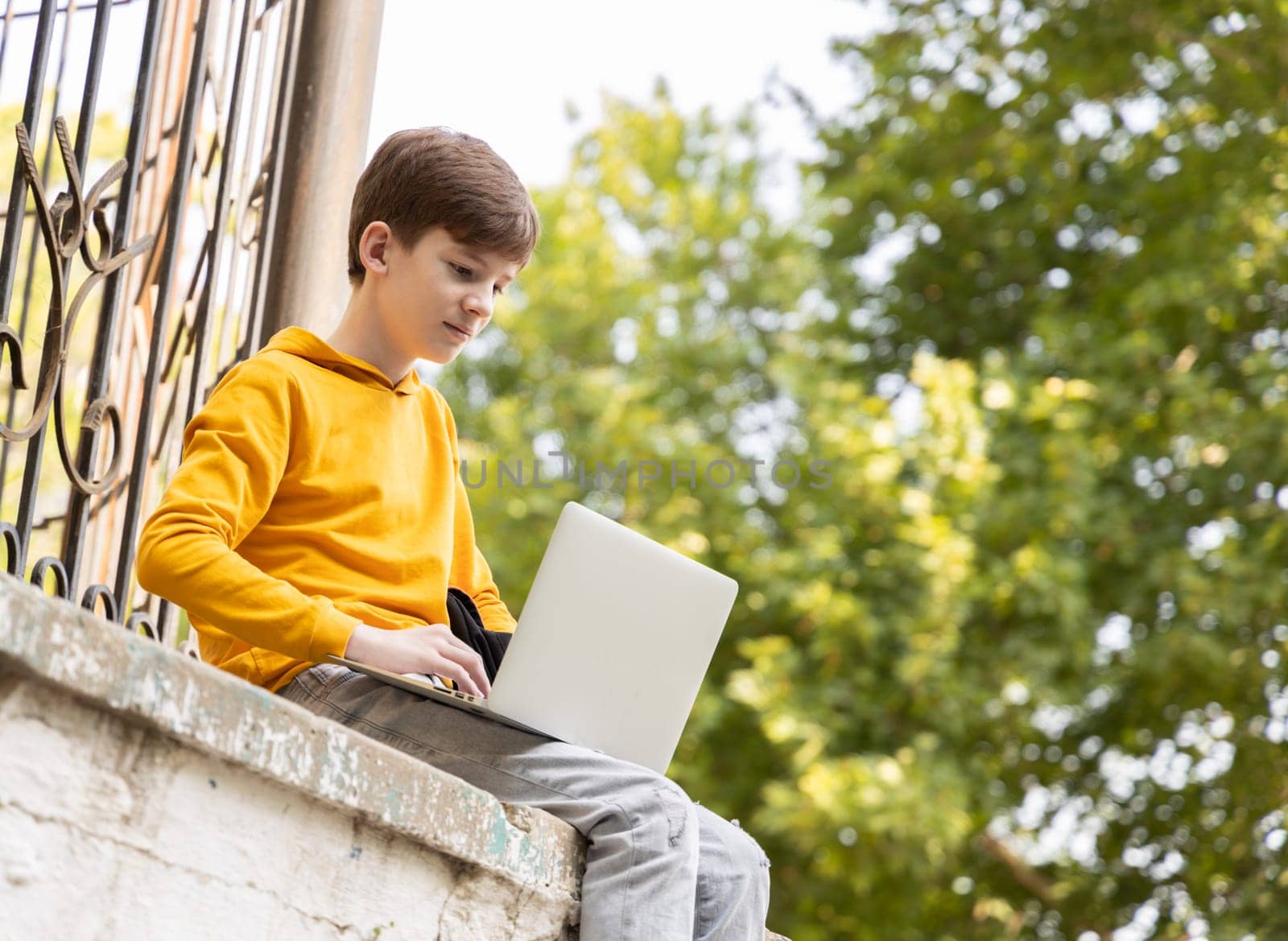 Caucasian child teen boy in yellow hoodie using laptop outdoors. Distant learning on the go. Coding, blogging, surfing, video call