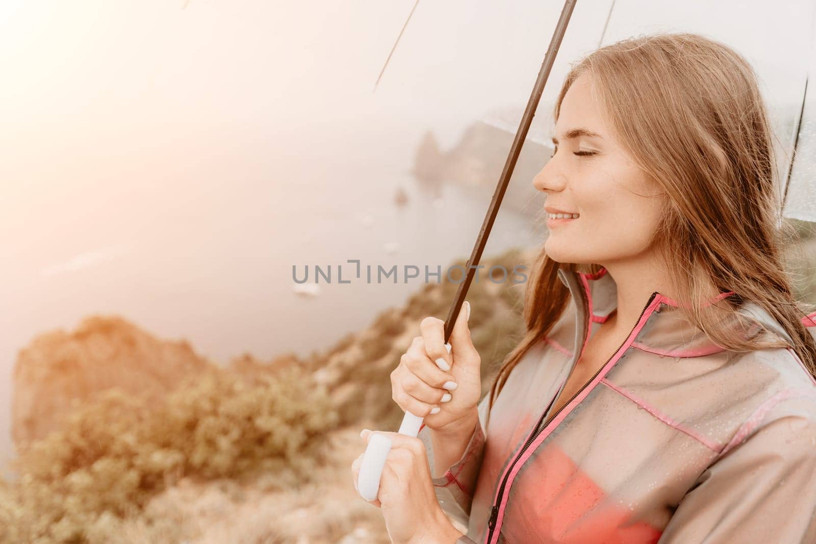 Woman rain park. Happy woman portrait wearing a raincoat with transparent umbrella outdoors on rainy day in park near sea. Girl on the nature on rainy overcast day