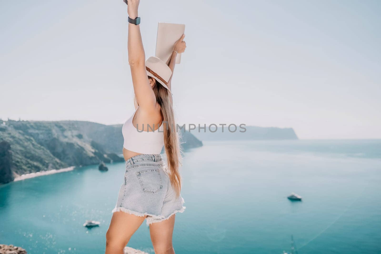 Woman sea laptop. Back view of happy young woman, female freelancer in straw hat working on laptop, keeping arms raised and cellebrating success while standing over blue sea beach. Work from anywhere. by panophotograph
