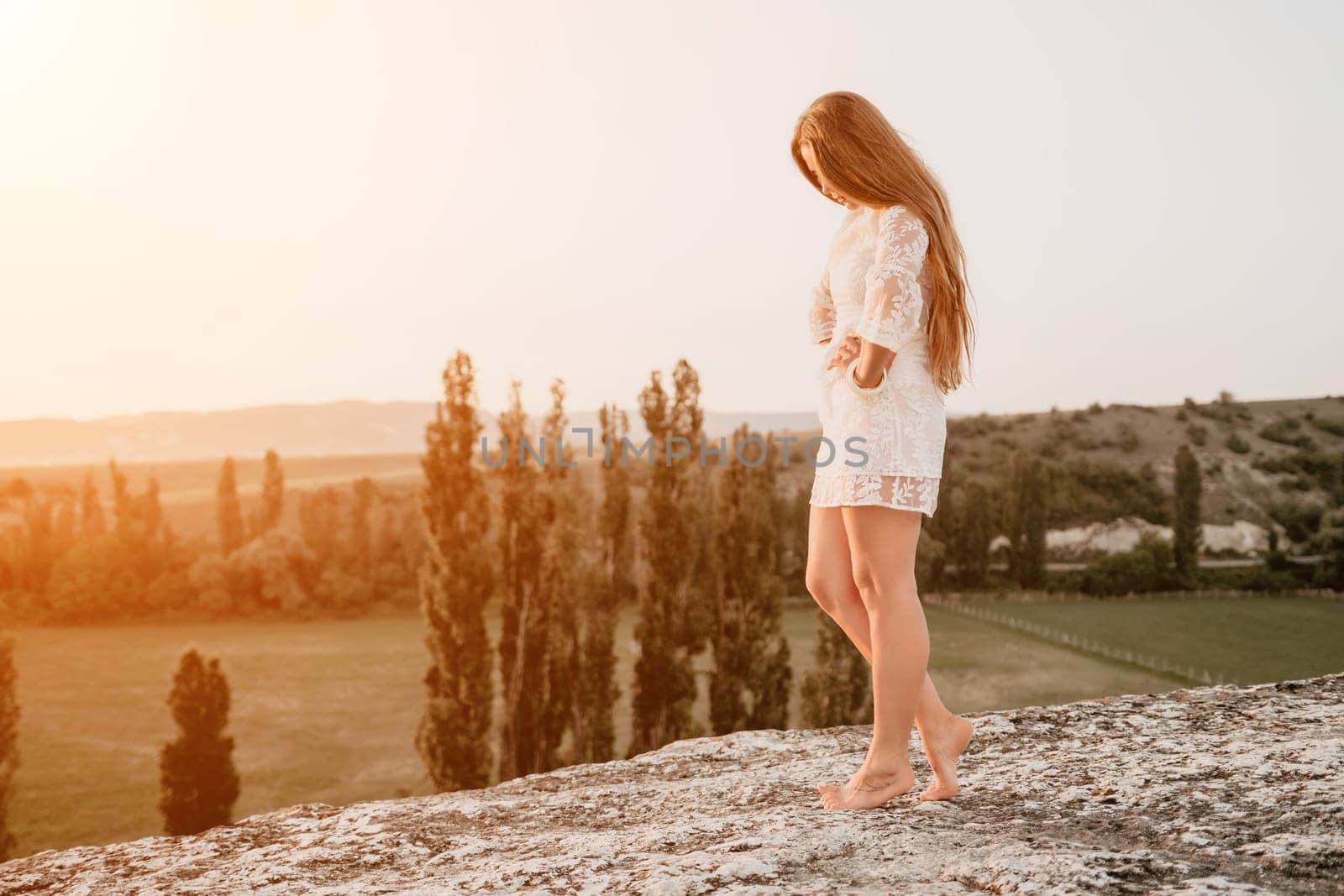 Happy woman in white boho dress on sunset in mountains. Romantic woman with long hair standing with her back on the sunset in nature in summer with open hands. Silhouette. Nature. Sunset. by panophotograph