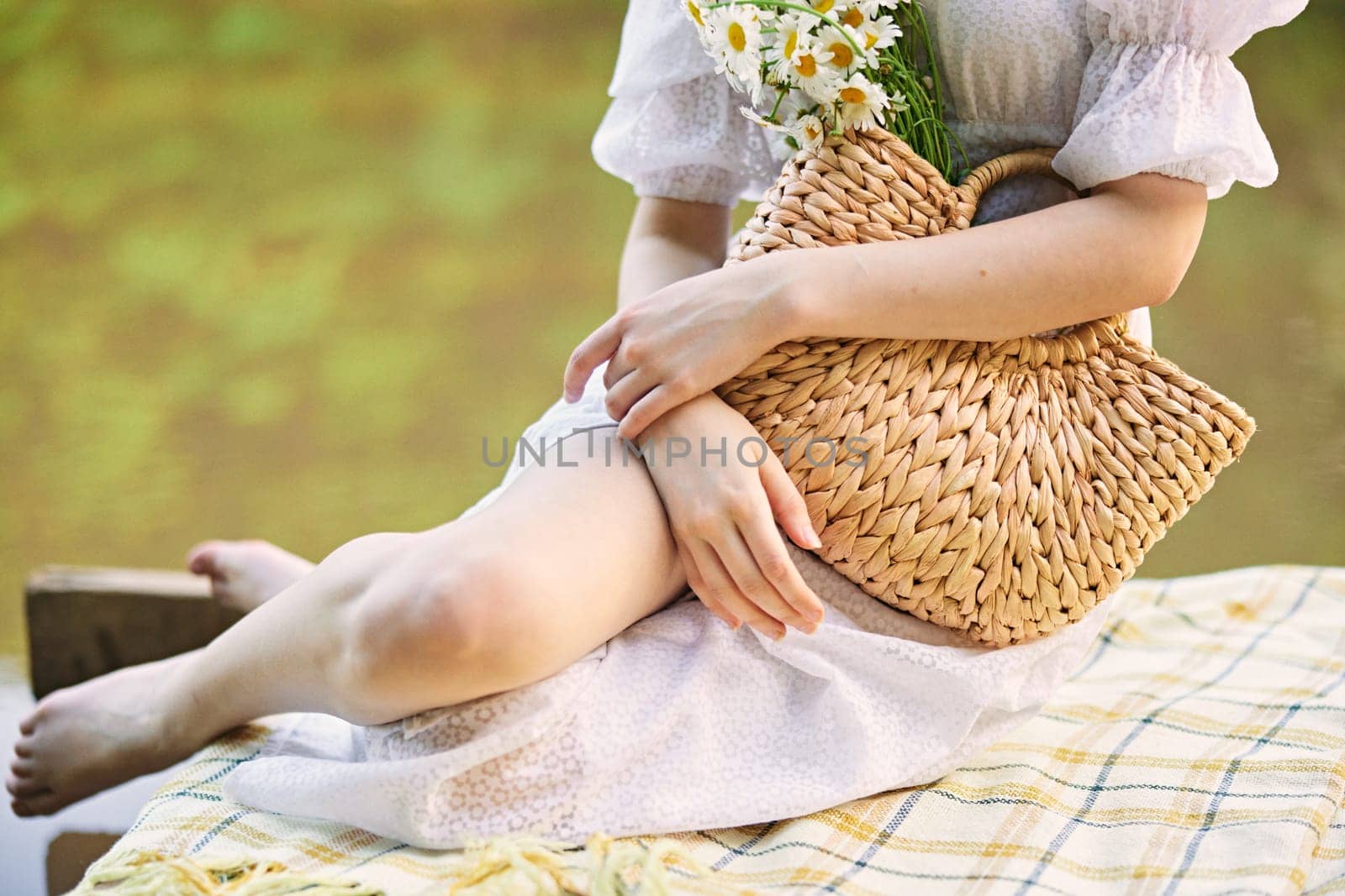 a woman in a light dress sits near the lake and holds a wicker basket with flowers in her hands. High quality photo