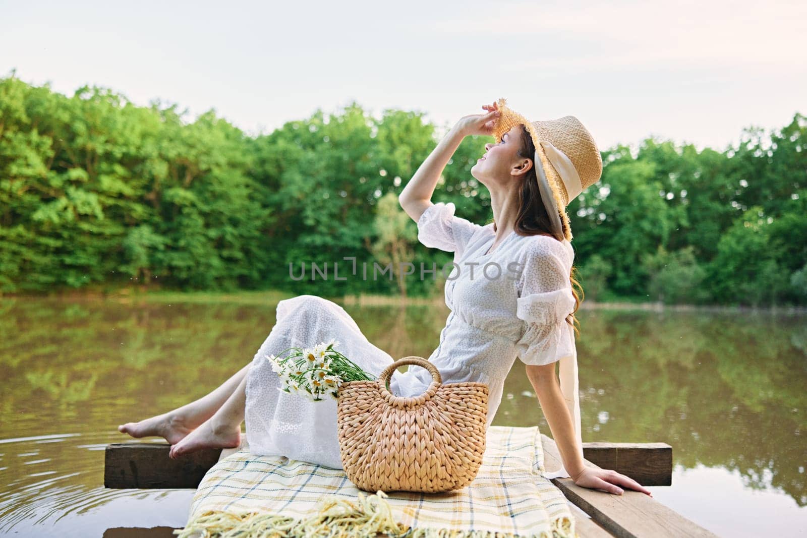 happy woman in a long light dress is resting by the lake sitting on a plaid and enjoying the view. High quality photo