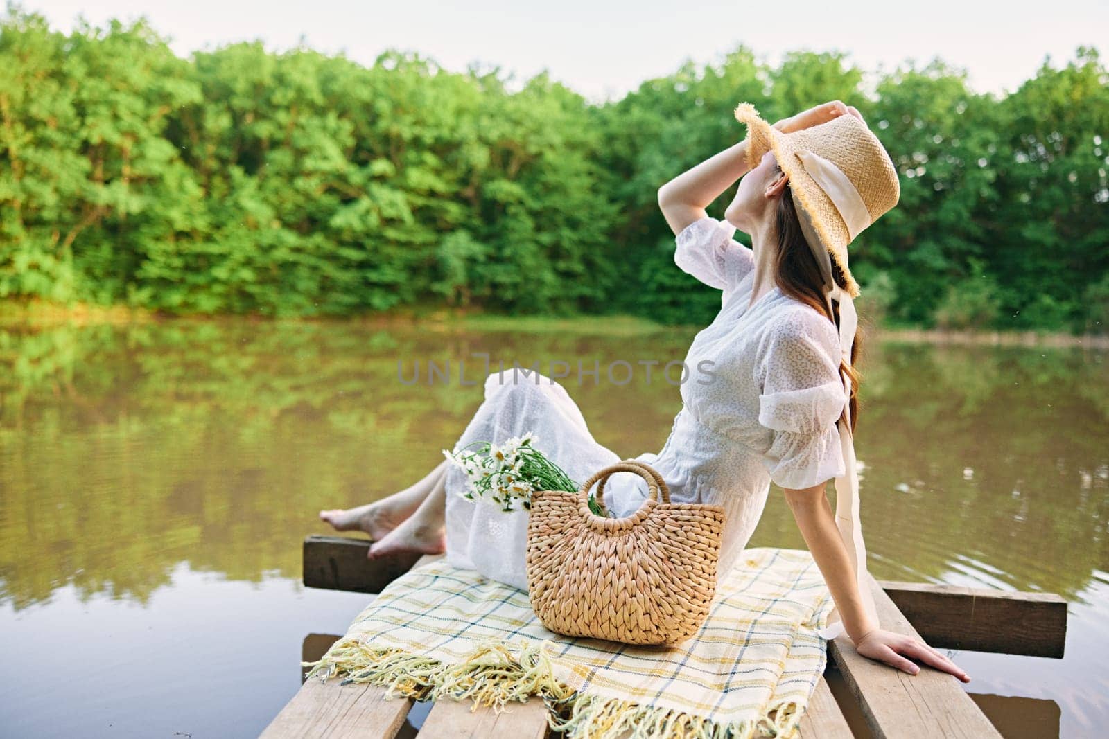 happy woman in a long light dress is resting by the lake sitting on a plaid and enjoying the view by Vichizh