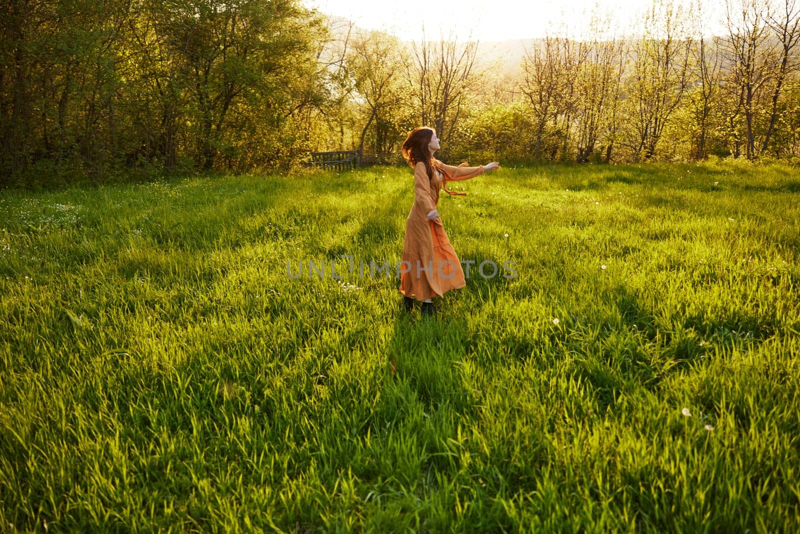 an attractive, slender, red-haired woman stands far away in a wide, green field during sunset in a long orange dress enjoying unity with nature and relaxation standing with her back to the camera by Vichizh