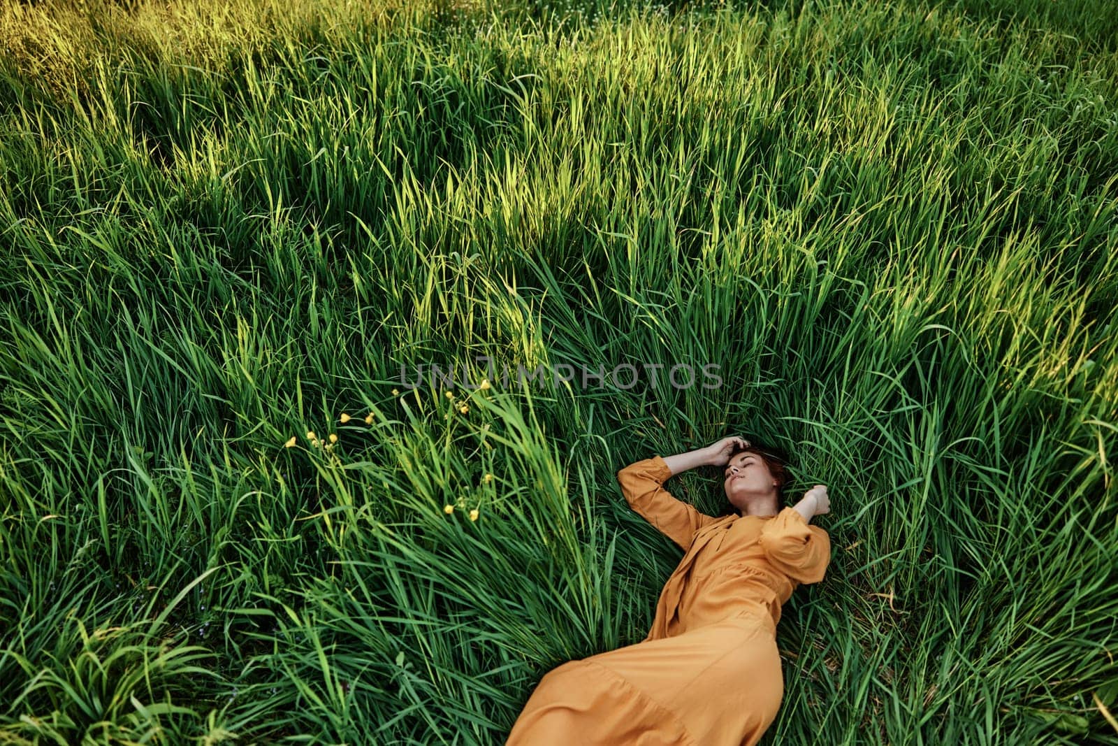 a sweet, calm woman in an orange dress lies in a green field with her hands under her head enjoying silence and peace. Horizontal photo taken from above by Vichizh