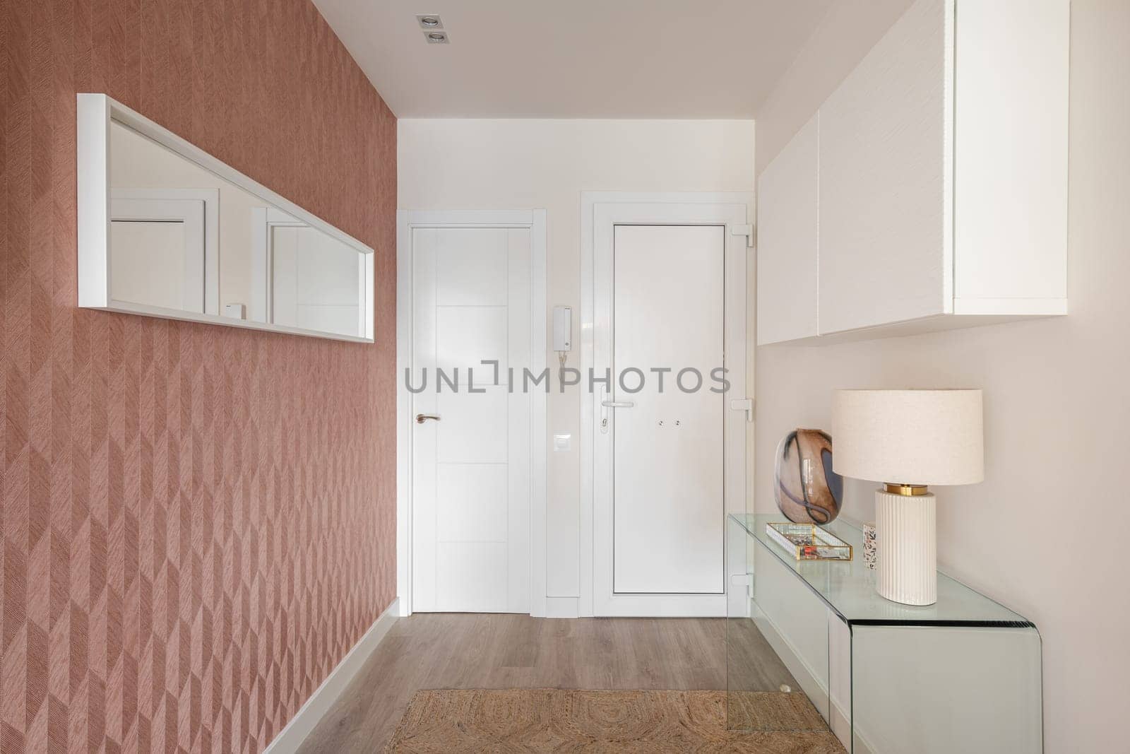 Cozy entrance hall in light colors with an intercom, glass console and wall cabinet with stylish light and an elongated mirror on a peach-colored wall. The concept of a simple laconic room design by apavlin