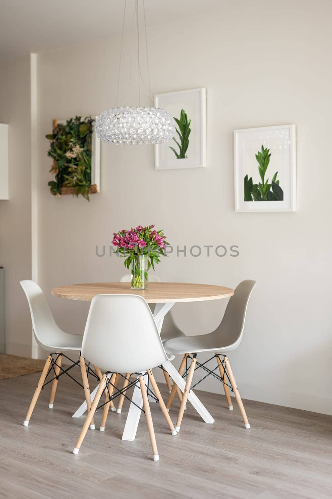 Vase with flowers stands on a wooden table next to chairs on background of wall with posters of green plants with beige wallpaper with a crystal chandelier. Botanical eco interior concept.