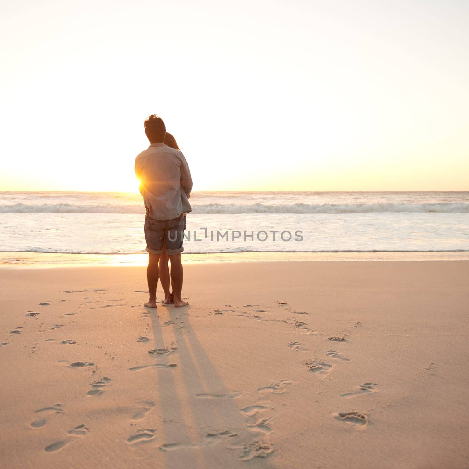 Love at sunset. Rearview shot of a young couple enjoying the sunset at the beach. by YuriArcurs