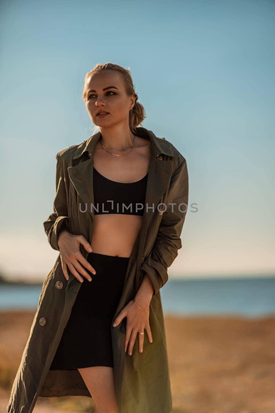 Portrait blonde sea cape. A calm young blonde in an unbuttoned khaki raincoat stands on the seashore, under the raincoat there is a black skirt and top by Matiunina