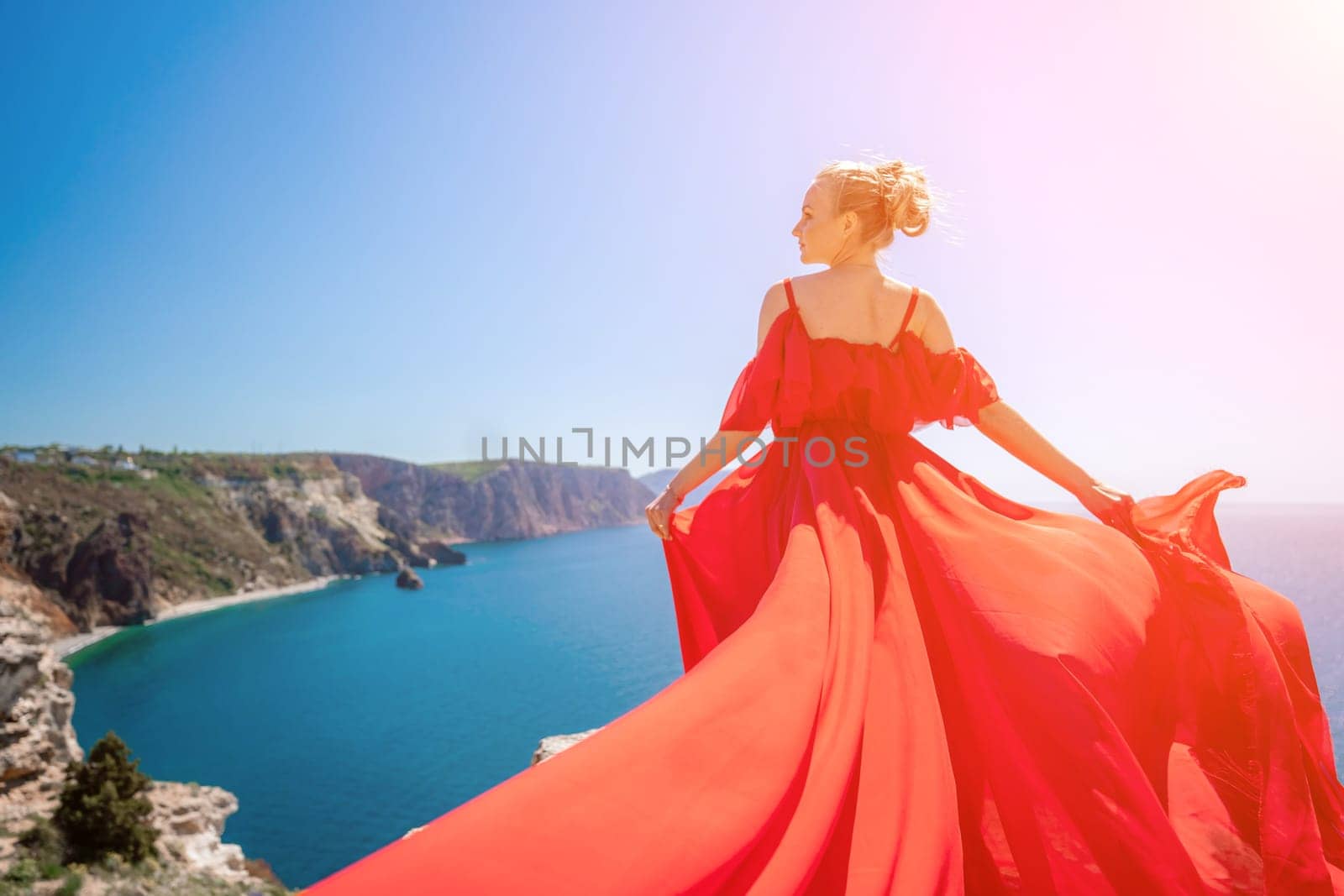 woman sea red dress. Blonde with long hair on a sunny seashore in a red flowing dress, back view, silk fabric waving in the wind. Against the backdrop of the blue sky and mountains on the seashore