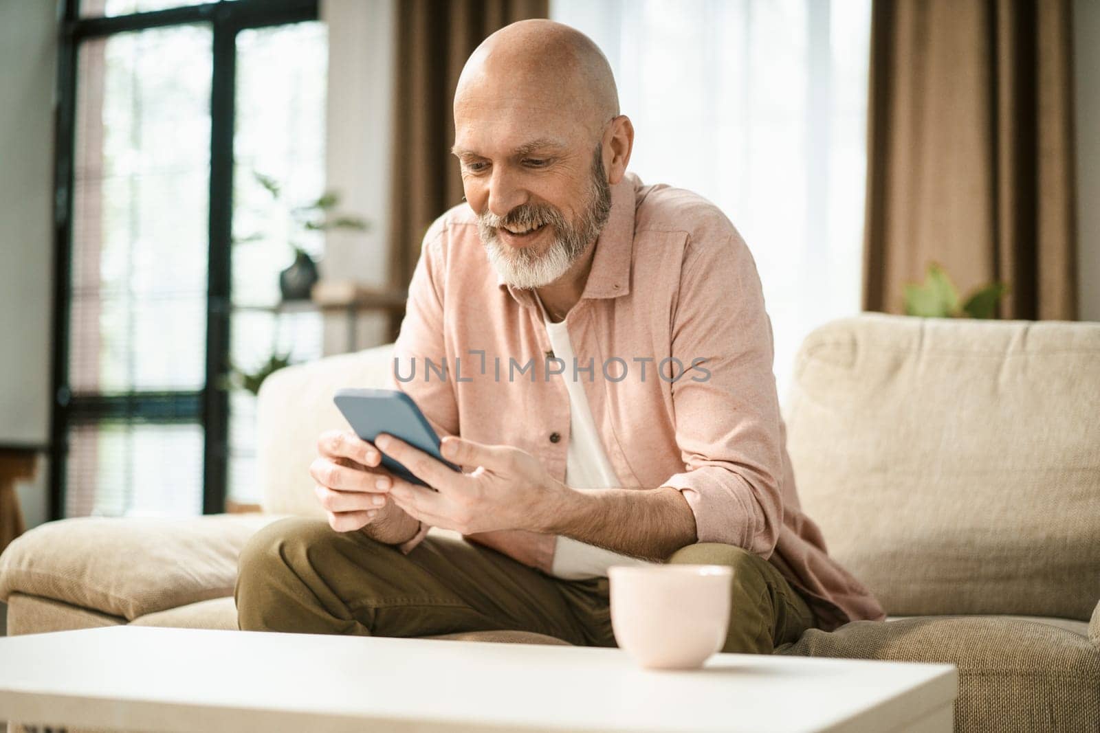 Happy mid aged man text message on phone while sit on sofa in home interior. by LipikStockMedia