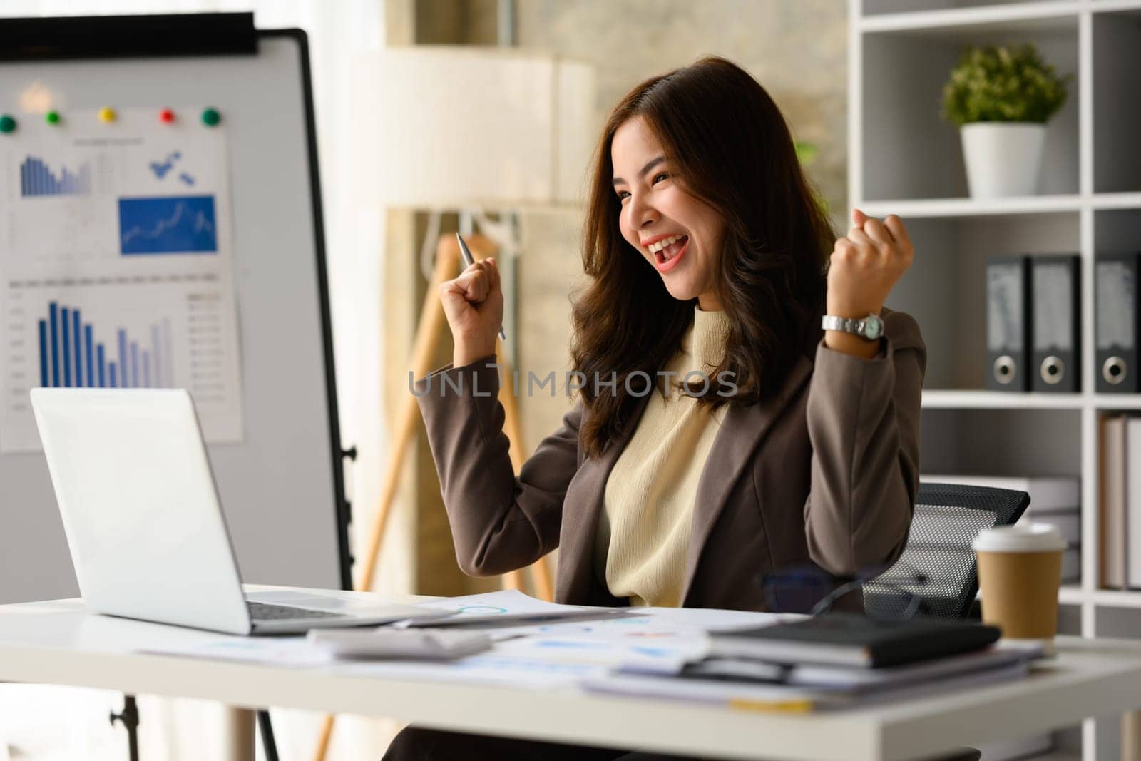 Surprised asian female entrepreneur looking at laptop screen, making winner yes gesture, celebrating business success by prathanchorruangsak