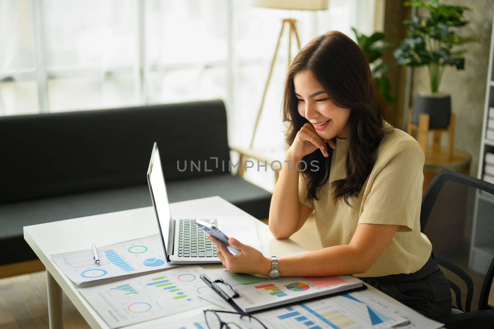 Charming young female entrepreneur smartphone at working desk with laptop and financial documents by prathanchorruangsak