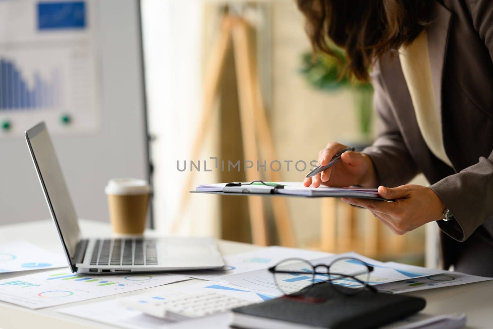 Cropped image of female investor holding clipboard and checking financial data on laptop computer by prathanchorruangsak