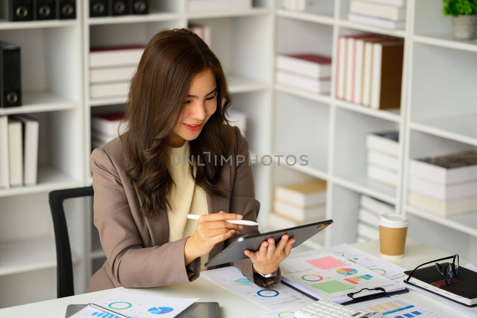 Beautiful female manager in stylish suit using digital tablet, searching online information at her workplace.