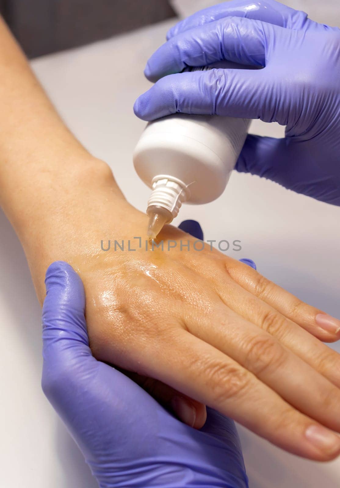 Manicure technician applies organic eco peeling on woman hand's skin from white tube. Nail master wears purple gloves. Nail hand spa treatment, care. Beauty procedure in process.Closeup.Vertical plane