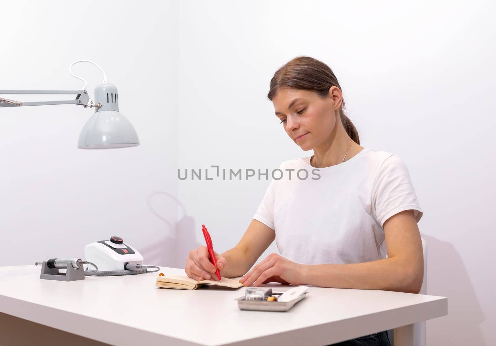 Beautiful nail beautician makes notes in notebook with pen, sitting at workstation, table in nail beauty treatment salon. Professional tools are on white table. Manicurist's routine. Horizontal