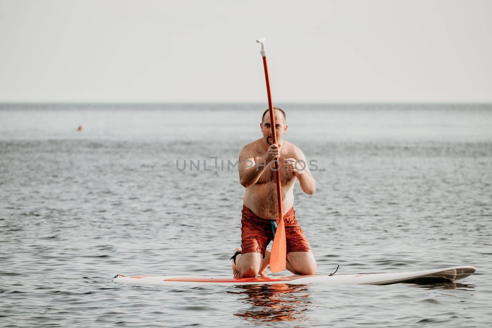 Active mature male paddler with his paddleboard and paddle on a sea at summer. Happy senior man stands with a SUP board. Stand up paddle boarding - outdor active recreation in nature