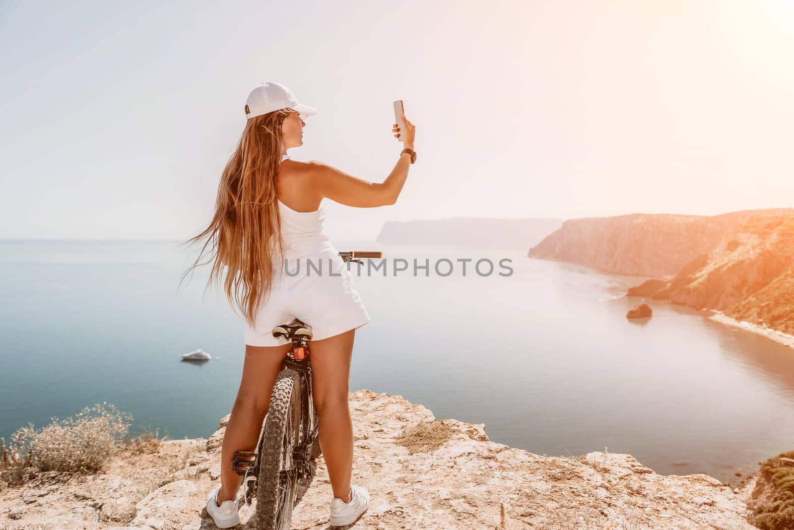 A woman cyclist on a mountain bike looking at the landscape of mountains and sea. Rear view of cyclist woman standing in front to the sea with outstretched arms. Freedom and healthy lifestyle concept