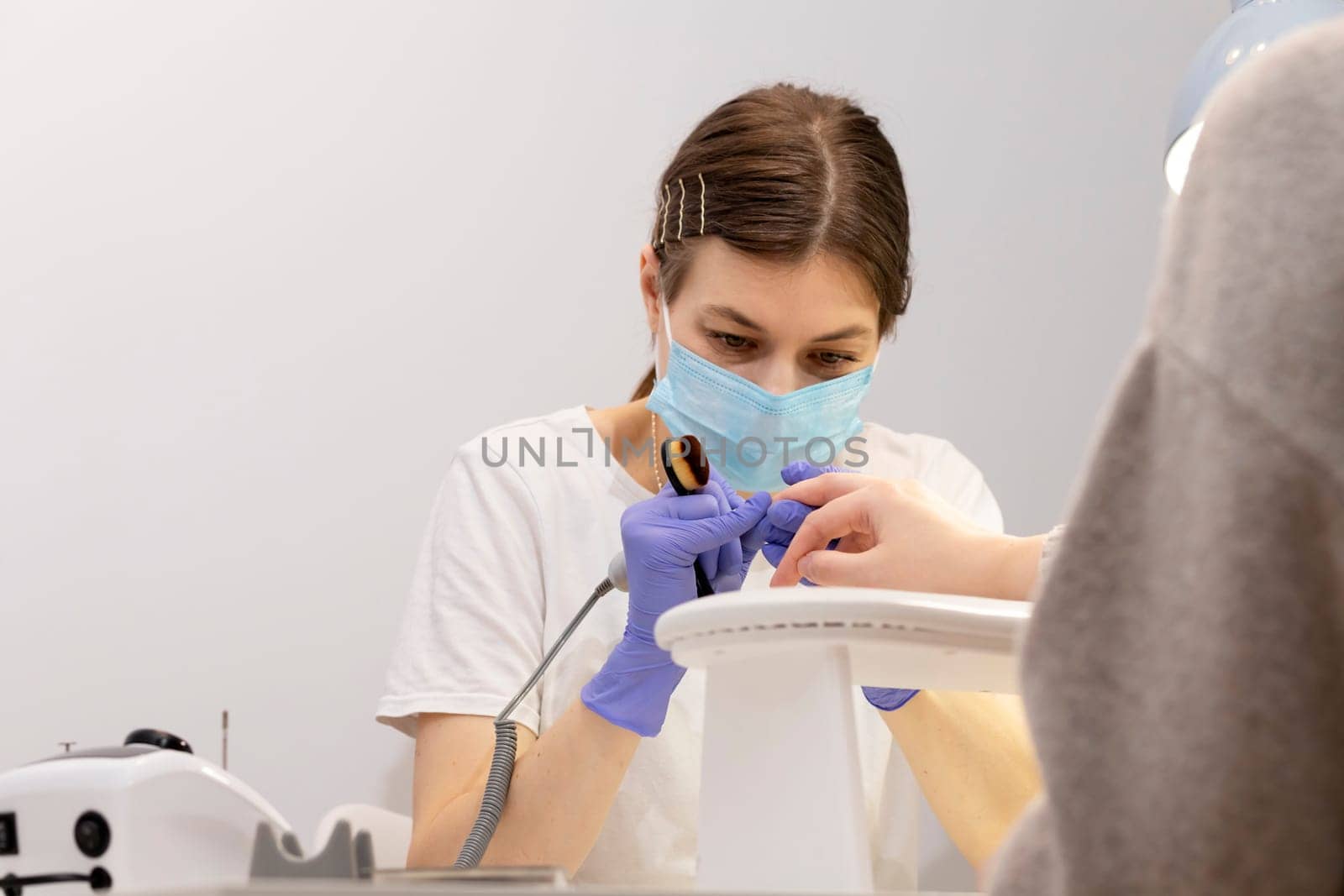 White nail technician in mask drills, removes client's cuticle with hardware machine, holding nail brush. Customer wears warm cloth, sweater. Cold season. Combined manicure, beauty treatment in spa.