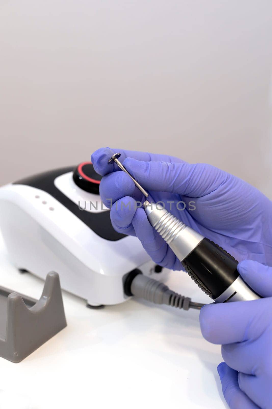 Nail beautician inserts nozzle in electric hardware drill file nail machine, wears purple groves. Nail treatment room in spa salon. Closeup, vertical. Manicure, pedicure concept. Copy space