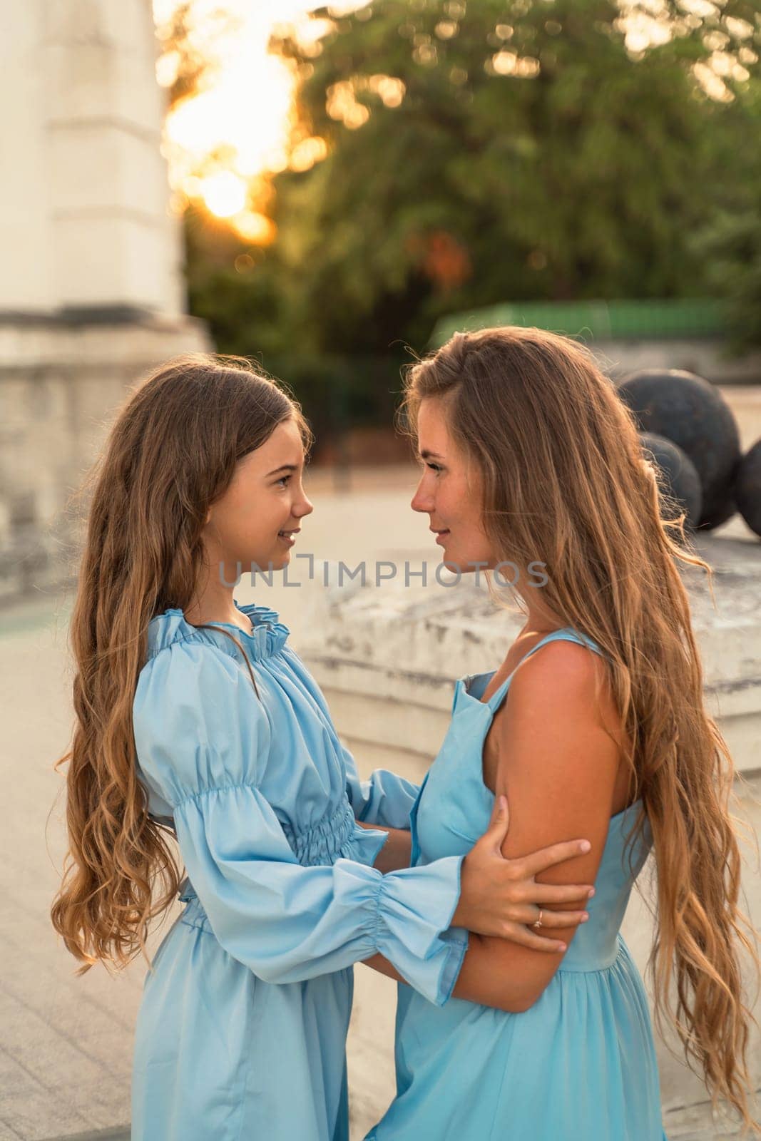 Portrait of a mother and daughter in blue dresses with flowing long hair against the backdrop of a sunset and a white building. They look at each other. Family stories on the weekend.