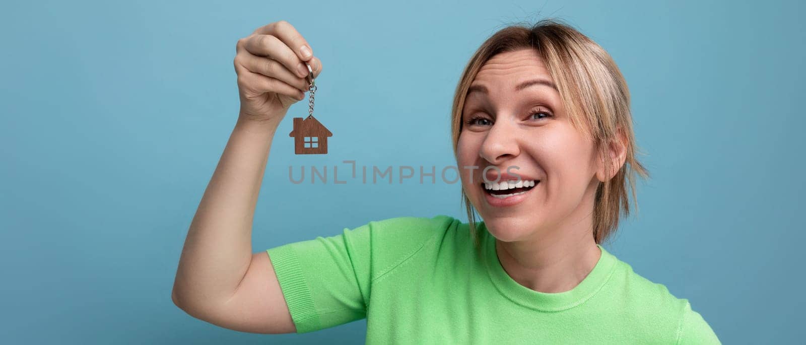 horizontal photo of the winner of a blond girl in a casual outfit who won the purchase of a home and holds a key ring on a blue background.