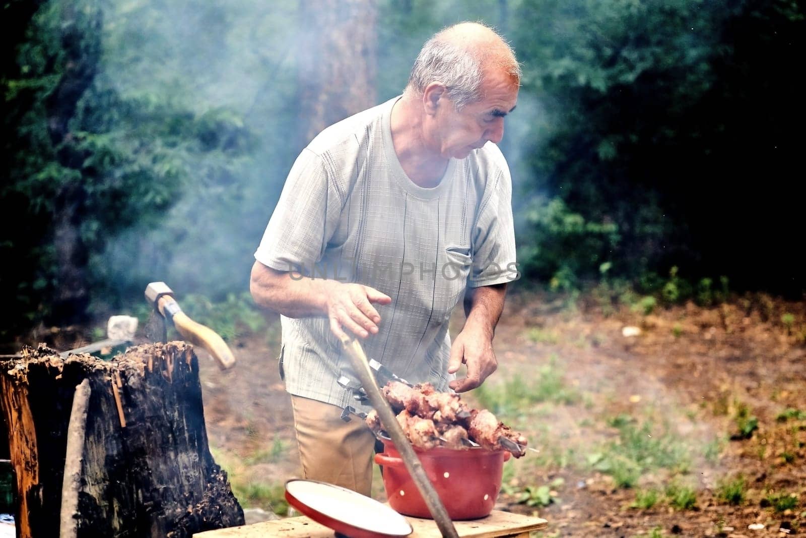 Elderly cheerful pensioner prepares meat for barbecue for cooking on grill fire by jovani68