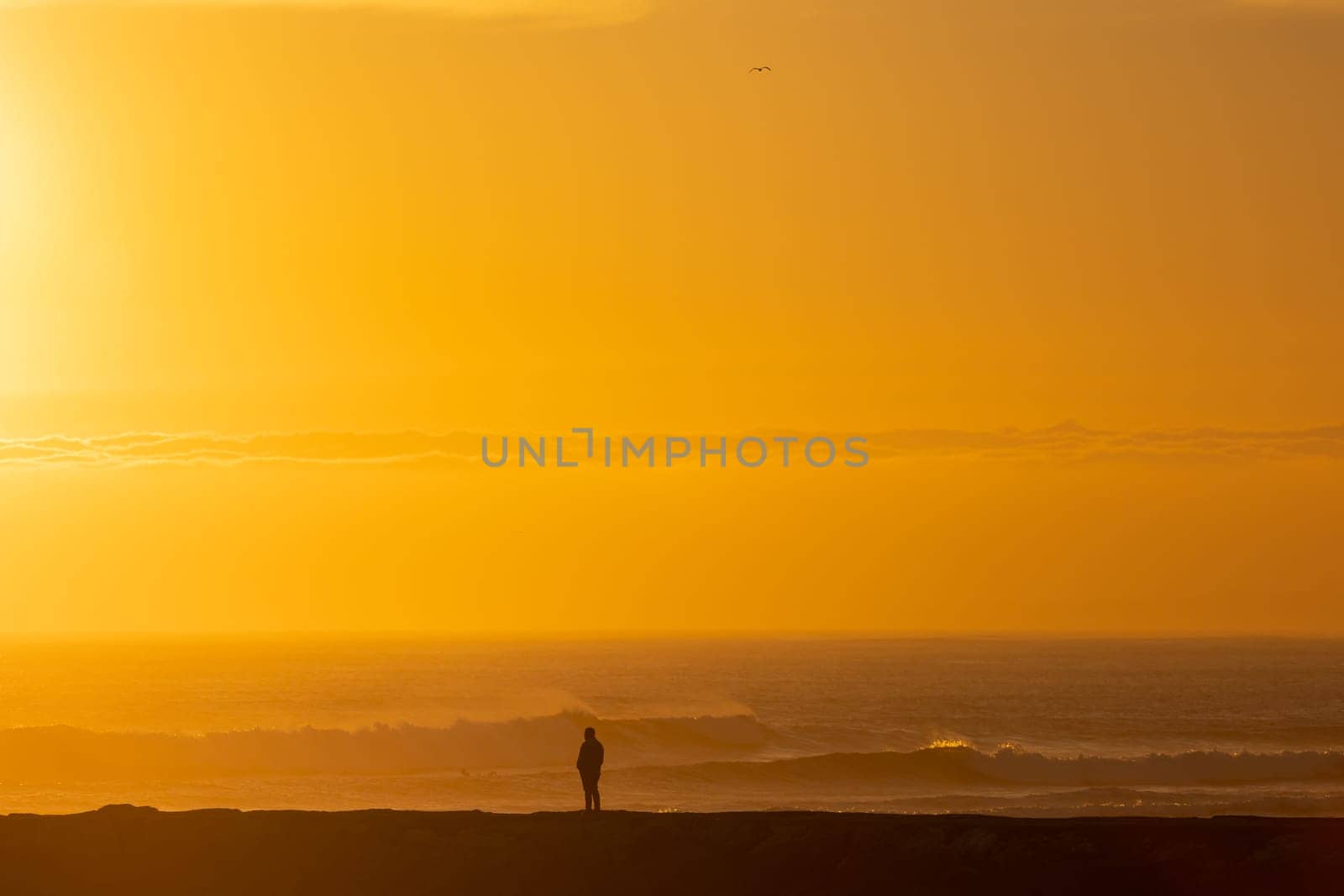 A lonely man stands by the sea at bright orange sunset. Mid shot