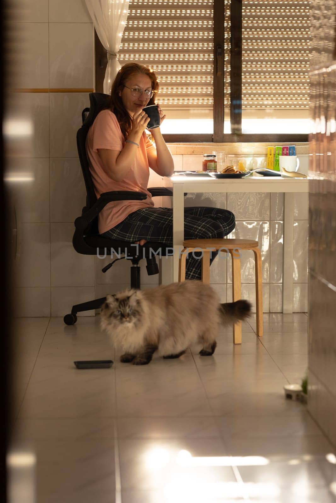 An adult woman sits at the kitchen table and drinks coffee - a cat sits next to her on the floor. Vertical shot